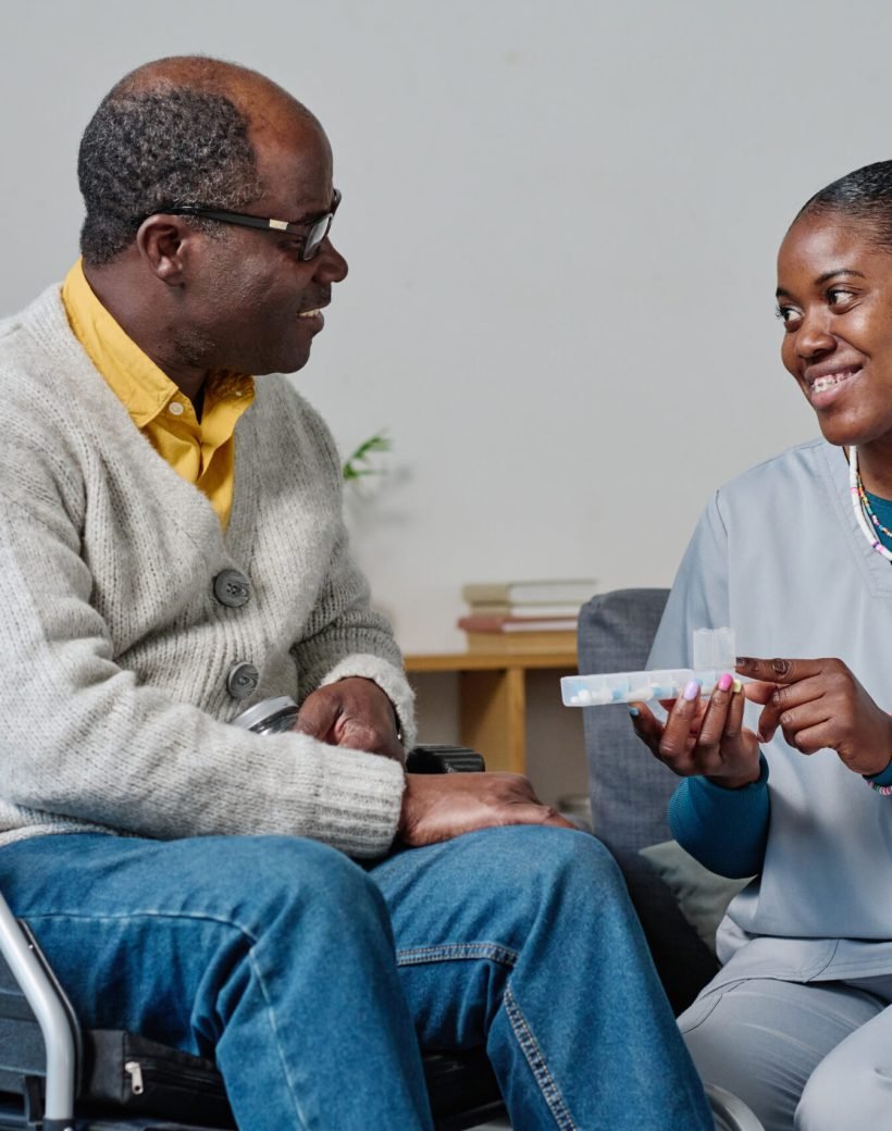 African caregiver showing organizer with pills to man with disability and explaining which pills to take while they sitting in room